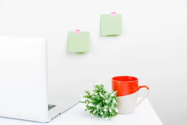 A white, tidy workspace setup with a laptop, sticky notes, a mug and a plant