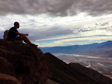 Arka planda dağlar olan bir uçurumda oturan bir adam.