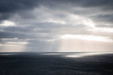 Kasvetli bir gökyüzünün altında büyüleyici bir deniz manzarası ve bulutlardan süzülen güneş ışığı.
