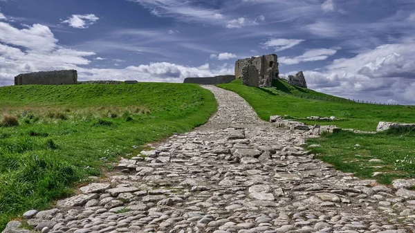 Narrow stone path through green valley leads to the ruins of Duffus Castle, located on grassy hill near Moray, Scotland, under blue cloudy sky