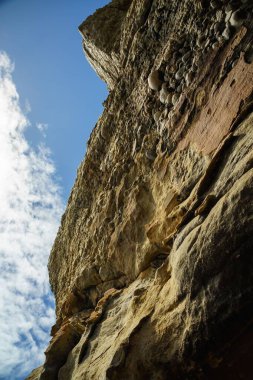 Açık mavi gökyüzünün altındaki yüksek kayalık bir uçurumun dikey görüntüsü.