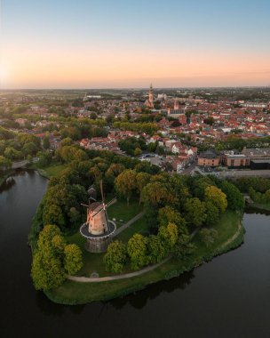 Yeşil sahildeki yel değirmeninin dikey hava görüntüsü ve arka planda şehir manzarası.