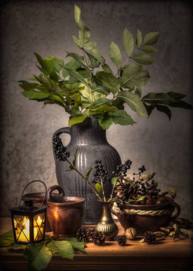 Bir kil sürahi, bir buket yaprak ve yanan bir fenerle, ahşap bir masada, durgun bir hayatın dikey görüntüsü.