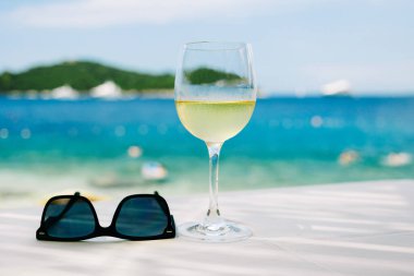 Bir bardak soğuk beyaz şarap ve arka planda plaj olan güneş gözlüğü.
