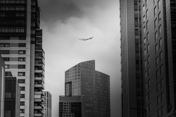 A grayscale of a plane flying high in the sky between modern skyscrapers