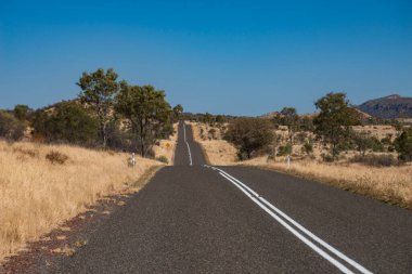 Mavi gökyüzü ve uzak dağları olan kırsal bir alanda düz bir yolun manzarası.