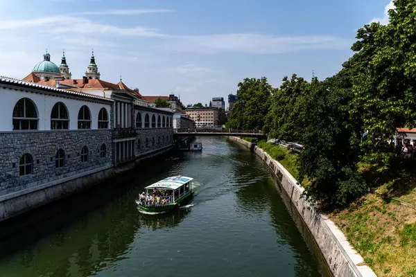 Ljubljana Nehri 'nin merkezinde, parlak bir güneş ışığı altında insanları taşıyan bir tekne görüntüsü.