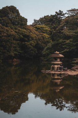 Heykelin yanında gölü ve gür ağaçları olan bir Japon bahçesinde oturan bir erkeğin dikey görüntüsü.