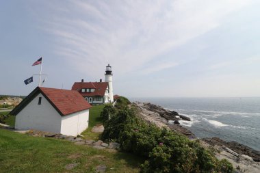 İnce bir gökyüzünün altındaki kayalık bir sahilde kırmızı çatılı evler ve deniz fenerinin doğal manzarası.