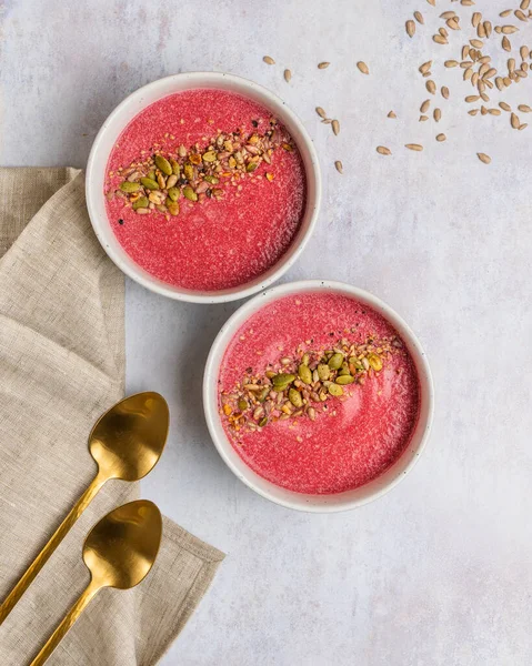 Altın kaşıklar ve peçetenin yanında beyaz pastel arka planda hindistan cevizi ve tohumlarla yapılmış pancar kreminin dikey görüntüsü..