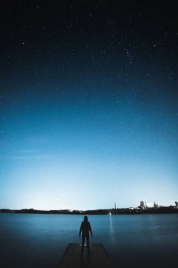 Göl kıyısındaki bir rıhtımda durup mavi yıldızlı gökyüzüne bakan birinin dikey görüntüsü.