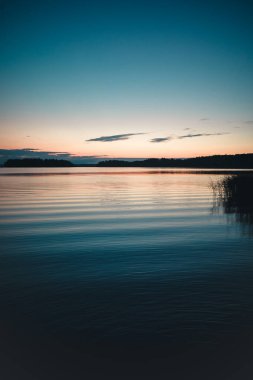 Gün batımında bitkilerle kaplı kayalık bir sahili yansıtan sakin bir gölün dikey görüntüsü.