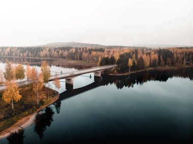Ormanlarla çevrili bir gölün sakin yüzeyine yansıyan bir köprünün hava görüntüsü.