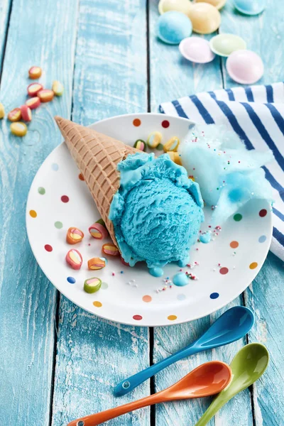A vertical of a blue bubble-flavored ice cream cone on a plate with colorful spoons and candies