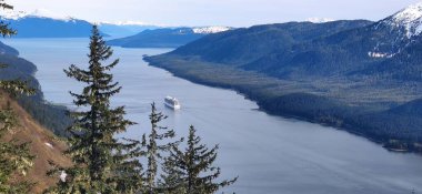 Güneşli bir günde, karlı dağlarla çevrili mavi bir gölde seyreden bir yolcu gemisinin kuş bakışı görüntüsü.