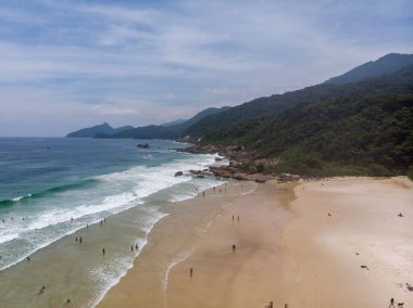 Deniz dalgalarına karşı yeşil tepelerle kaplı kumlu bir plajda dinlenen insanların hava görüntüsü.