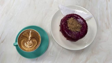 Bir fincan kahvenin yüksek açılı görüntüsü latte sanatıyla ve ahşap yüzeyde mor jöleli kek.