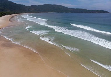 Yeşil tepelerle kaplı kumlu bir sahile çarpan deniz dalgalarının hava görüntüsü.