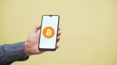 Cep telefonu taşıyan bir elin yakın plan fotoğrafı ve üzerinde beyaz bir arkaplanda izole edilmiş bitcoin logosu var.