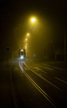 Gece sisli bir caddede ilerleyen tramvayın dikey görüntüsü.