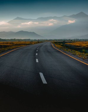 Renkli tarlalar ve bulutlu gökyüzüne karşı dağlarla çevrili bir yol.