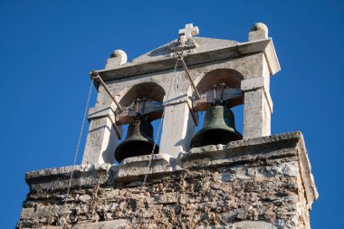 Taştan bir haçın alçak açılı görüntüsü ve parlak güneş ışığında mavi gökyüzüne karşı eski bir kilisenin iki çanı.