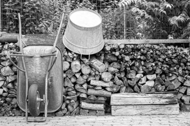 Eski bir kova ve arka planda yakacak odunları olan eski bir el arabasının gri tonlu görüntüsü.