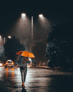 Zor bir günün ardından, yağmurlu bir gecede sarı şemsiyeli bir adamın eve yürüdüğü dikey bir fotoğraf.
