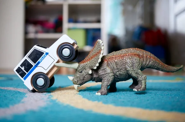 A closeup of a Triceratops Dinosaur toy pushing a toy car on a blue rug
