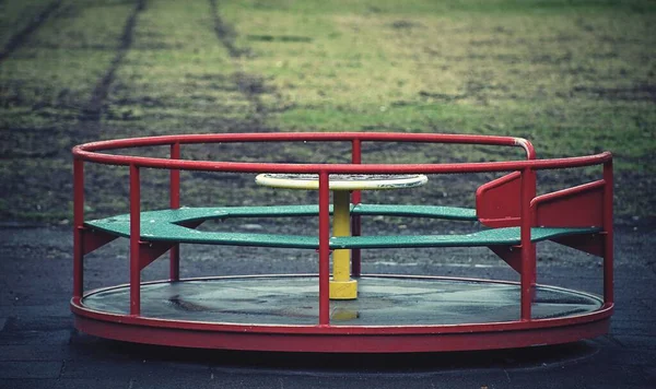 緑と赤の回転するカルーセルが一人で遊び場に立っていて 背景には土と草があります — ストック写真