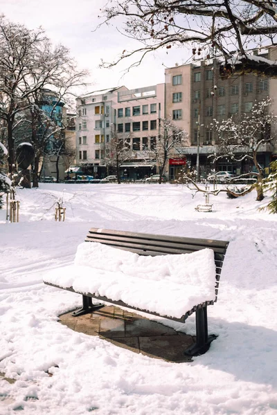 A snow-covered bench in a park with a view of apartment buildings