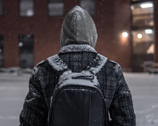 一个头罩里的男人穿着一件背着背包的外套站在雪夜的背影 — 图库照片
