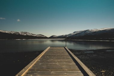 Kışın dağlık bir arka planda, gölün kıyısındaki ahşap bir iskelenin manzarası.