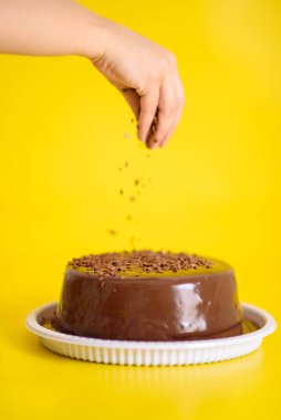 Çikolatalı kekin üzerine çikolata kırıntıları düşüren bir elin dikey görüntüsü, sarı arka planda izole edilmiş, pişirme dersleri için.