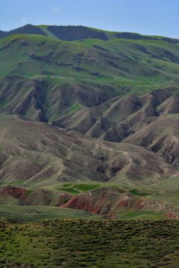 Mavi gökyüzünün altındaki yeşil dağların altındaki yüksek bir tepenin dikey görüntüsü.