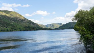 Mavi gökyüzünün altındaki yeşil tepelerin altında güzel bir göl suyu manzarası.