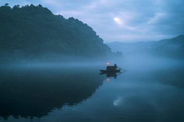 Gün batımında sisli gölün yüzeyine yansıyan ışık ile küçük balıkçı teknesinin gölünün gölgesinin görsel bir görüntüsü.