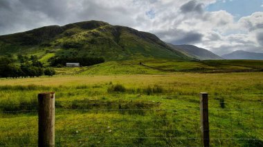 Bulutlu bir gökyüzünün altındaki dağlar ve ön planda bir çit