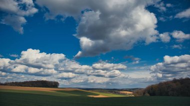 Bulutlu mavi gökyüzünün altındaki ağaçlarla kaplı yeşil bir arazi manzarası.