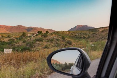 Çimen tarlaları ve tepeleri olan kırsal bir arazinin dikiz aynasından sürücünün görüntüsü.