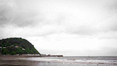 Gri gökyüzünün arka planında yeşil bir tepe olan boş bir sahil..