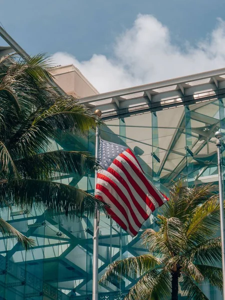 A vertical shot of US Flag against palm trees and a modern building