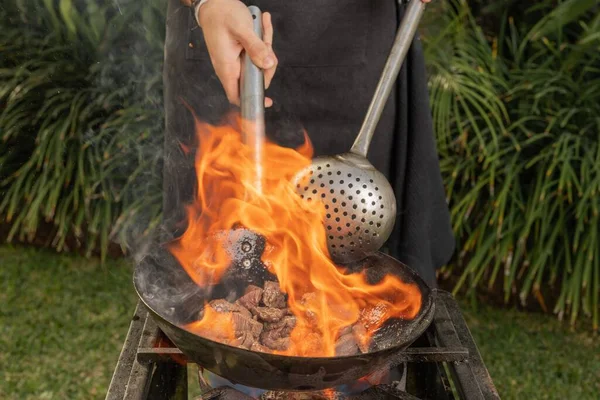 Cropped photo of a cook flaming chunks of meat in a frying pan with fire in the middle of a garden