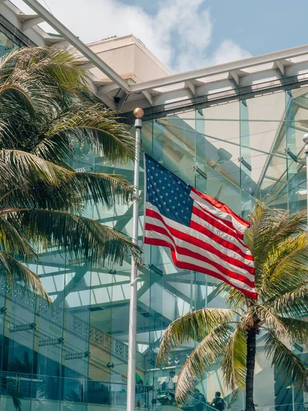 A vertical shot of US Flag against palm trees and a modern building