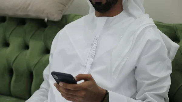 Арабський Чоловік Одягнений Білу Істоту Пробирається Телефону Сидячи Зеленому Дивані — стокове фото