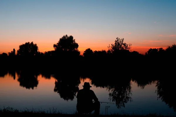 日没時に湖の海岸に座って後ろから漁師のシルエット — ストック写真