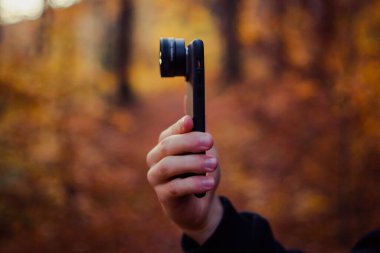 Sonbahar ormanında kameralı bir lensle telefonu tutan bir el.