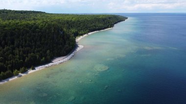 Mavi denize karşı yeşilliklerle kaplı kumlu bir sahilin kuş bakışı görüntüsü.