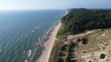 Mavi denize karşı yeşilliklerle kaplı kumlu bir sahilin kuş bakışı görüntüsü.