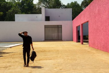 A fashionable man dressed in black with a backpack standing by an architectural work of Luis Barragan clipart